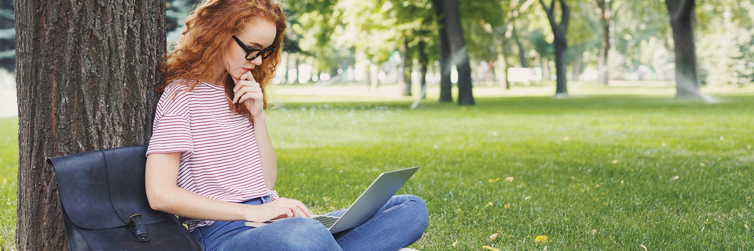 Student looking at laptop and sitting under tree